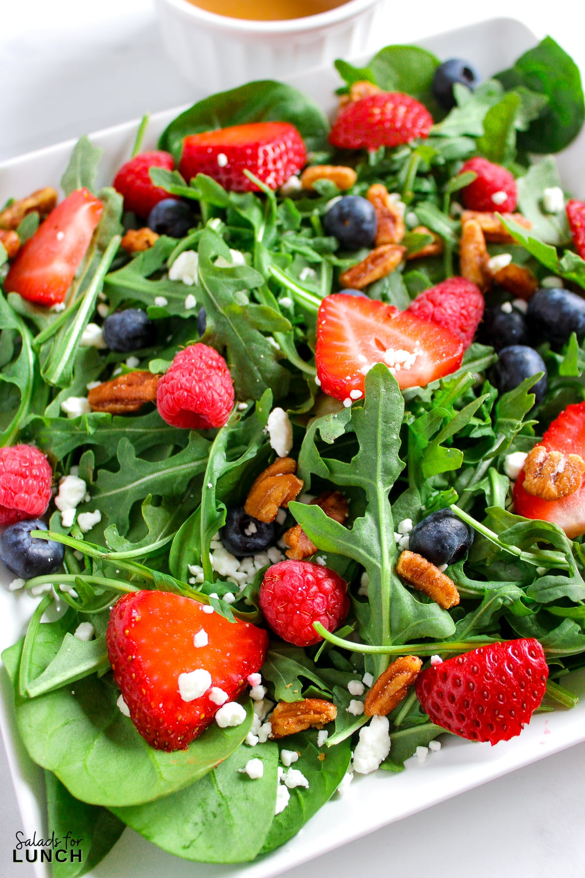 A close up photo of a salad with arugula,berries and honey roasted pecans topped with a creamy balsamic vinaigrette on a plate.